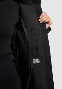 Zwarte zip-up jas met een zachte buitenstof en een glanzende binnenkant. Bevat een zijzak met rits en een label met de tekst "Khujo™ originele wisseldraden."
