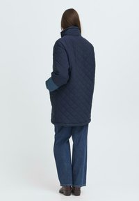 Chaqueta acolchada de color marino con cuello alto, que presenta mangas largas y un corte relajado. Combinada con jeans de pierna ancha azules y zapatos oscuros.