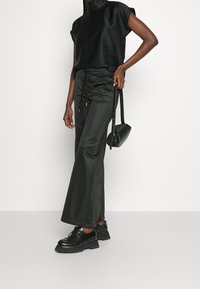 Mujer vistiendo pantalones anchos negros, top sin mangas negro, zapatos negros toscos, sosteniendo un pequeño bolso negro contra un fondo liso.