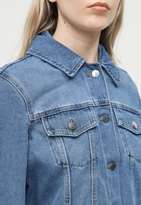 Close-up af en person iført en blå denimjakke med knappede brystlommer og en klassisk krave, der viser en del af halsen og den nederste del af ansigtet.