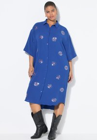 Robe bleu à boutons, en tissu texturé, ornée de motifs colorés de rayons de soleil brodés, avec des manches amples. Bottes de cowboy noires.