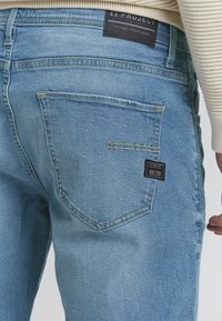 Jeans de mezclilla azul claro con un bolsillo trasero que muestra una etiqueta negra, con texto blanco, detalles de costura y un ligero efecto desgastado.