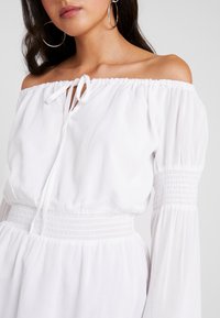 Robe blanche à épaules dénudées avec un col à cordon, taille smockée et manches longues amples. Tissu doux avec une texture lisse.