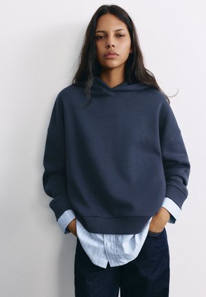 Femme aux longs cheveux foncés porte un sweat à capuche bleu marine sur une chemise à rayures bleu clair et un pantalon foncé, debout les mains dans les poches contre un mur blanc.