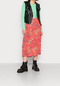 Un gilet en cuir noir sur un haut vert menthe, associé à une jupe midi à fleurs corail et des bottines noires à lacets avec des semelles épaisses.