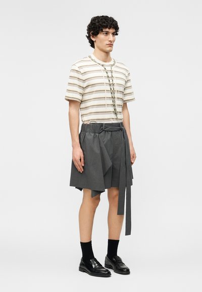 Giovane con capelli ricci che indossa una camicia beige a righe a maniche corte, pantaloncini grigi a gamba larga con cintura, calzini neri e mocassini neri, in piedi.