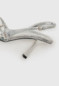 Stříbrná kovová vysoká sanda s páskovým designem, nastavitelným kotníkovým sponou a elegantním stiletto podpatkem s černým hrotem.