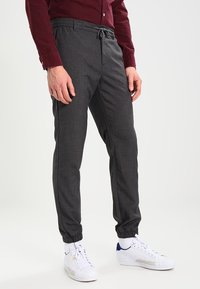 Uomo in pantaloni jogger grigi, sneakers bianche e camicia button-up bordeaux che si trova davanti a uno sfondo neutro.