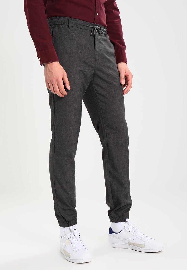 Uomo in pantaloni jogger grigi, sneakers bianche e camicia button-up bordeaux che si trova davanti a uno sfondo neutro.
