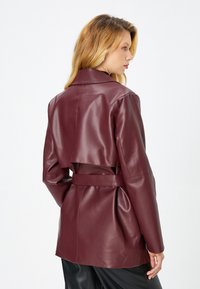 Burgunderfarbene Kunstlederjacke mit tailliertem Design, hohem Kragen und Ausschnitt im Rücken. Glatte Textur und maßgeschneiderte Passform.