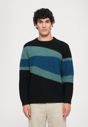 Mann mit lockigem, dunklem Haar, der einen schwarz-blau-grünen gestreiften Strickpullover und hellbeige Hosen trägt, steht vor einem schlichten Hintergrund.