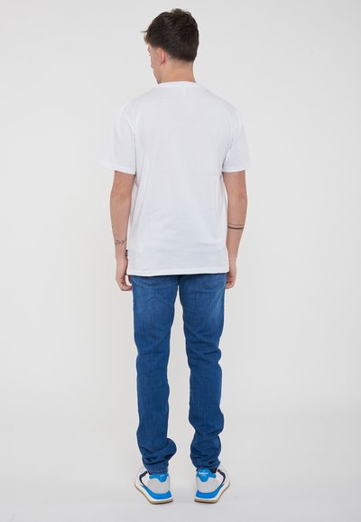 T-shirt bianca in cotone, colletto tondo, vista posteriore. Abbinata a jeans in denim blu. Le scarpe presentano dettagli grigi, bianchi e blu. Design minimalista.