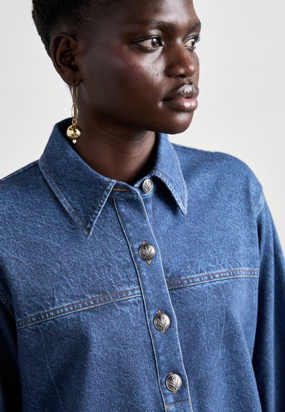 Denim shirt with light blue color, classic collar, silver buttons, and orange stitching details. Smooth texture with a slightly loose fit.