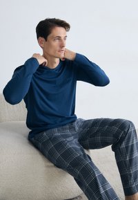 Blauwe langetermijnshirt van gladde stof, gecombineerd met grijze geruite pyjamabroek met een geblokt patroon. Zachte achtergrondtextuur.