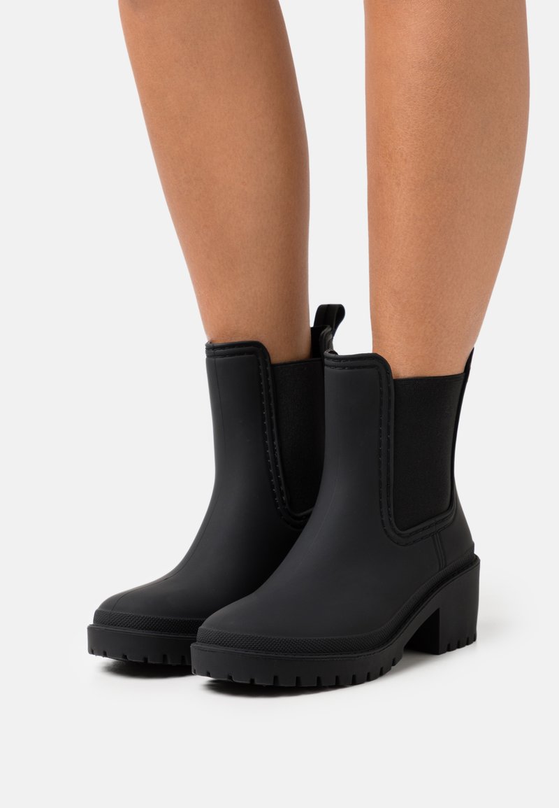 Botines negros con un exterior de goma, paneles laterales elásticos y un tacón grueso. Suela texturizada para un buen agarre y pestaña de tirón para facilitar el uso.