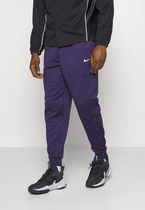 Mann trägt lila Nike-Jogginghosen mit Seitentaschen und schwarze und weiße Nike-Sneaker steht vor einem einfarbigen Hintergrund.