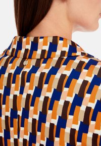 Bunt gemusterter Stoff mit orangefarbenen, blauen, braunen und beigen abstrakten Formen. Der Kragen hat eine Knopfdetail auf der Rückseite.