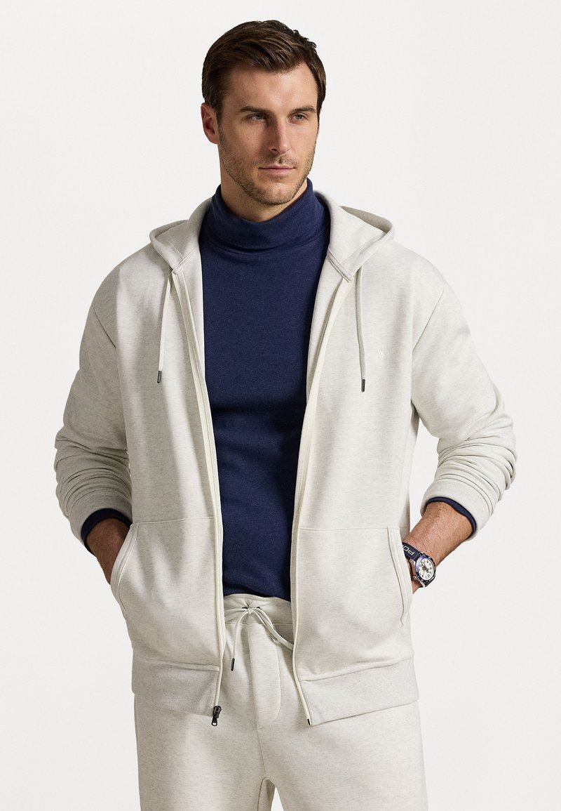 Light gray zip-up hoodie with drawstrings, featuring ribbed cuffs and pockets, worn over a dark blue turtleneck, paired with matching sweatpants.