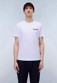 Bílý bavlněný tričko s kulatým výstřihem, na levé části hrudi malým barevným logem a krátkými rukávy, v kombinaci s tmavými kalhotami.