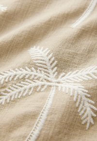 Tessuto di lino in tonalità beige con motivi di felce ricamati in bianco. La trama appare morbida con cuciture visibili. Estetica delicata e naturale.