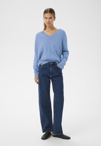 Világoskék V-nyakú pulóver sötétkék, magas derekú, egyenes szárú farmerral és fekete slip-on cipővel párosítva. Egyszerű, laza fazon.