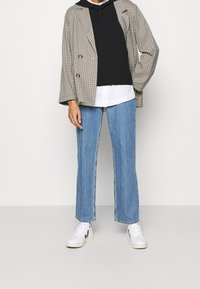 Blazer oversize en noir et blanc à motif pied-de-poule, superposé sur un sweat à capuche noir. Un jean droit bleu clair et des baskets blanches complètent la tenue.