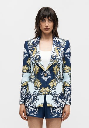 Γυναίκα που φοράει μπλε σκούρο και ανοιχτό μπλε σακάκι και αντίστοιχα σορτς με χρυσά και λευκά περίτεχνα λουλουδένια σχέδια, στέκεται μπροστά σε απλό φόντο.