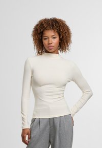 Langärmliges, cremefarbenes Rollkragenshirt mit taillierter Passform und glattem, dehnbarem Material. Kombiniert mit grauen, locker sitzenden Hosen.