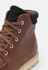 Bottines en cuir brun avec une surface texturée, col en suede noir, œillets en métal et lacets plats bruns ; semelle en caoutchouc avec un motif de crampons.