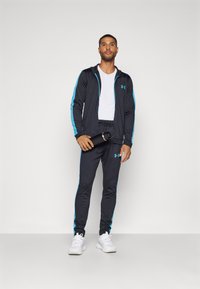 Survêtement de sport noir avec accents latéraux bleus, veste zippée, pantalon ajusté assorti, t-shirt blanc en dessous, et une bouteille d'eau à la main.