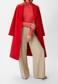 Een vrouw in een felrode jas over een koraalkleurige turtle neck en crème hoge tailles, wijde broek tegen een effen achtergrond.