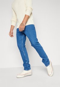 Pánské světle modré džíny z denimu s úzkým střihem, viditelným šit ím a hladkou texturou, doplněné bílými teniskami a krémovým svetrem.