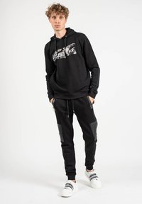 Sudadera con capucha negra con gráfico en la parte frontal, combinada con pantalones deportivos negros con acentos texturizados y bolsillos con cremallera, llevada con zapatillas blancas.