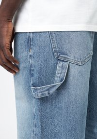 Jean en denim bleu clair avec une poche latérale à rabat, des détails de piqûres visibles, et une coupe décontractée. La main est placée près de la poche.