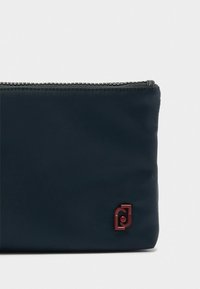 Marineblå nylonpung med en struktureret overflade, der har et kontrastfarvet bordeaux-logo og en lynlåslukning på toppen. Dimensioner er ikke synlige.