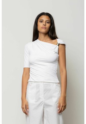Giovane donna con lunghi capelli scuri che indossa un top bianco asimmetrico con dettaglio metallico e pantaloni bianchi, in piedi davanti a uno sfondo chiaro.