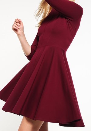 Nainen, jolla on yllään liehuva, polvipituinen viininpunainen mekko, jossa on kolmanneksen pituiset hihat, pyörähdyksessä kuvattuna yksinkertaisen taustan edessä.