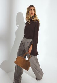 Braune Wildlederhandtasche mit kurzen Griffen, kombiniert mit einem dunklen Oberteil und hellgrauen, weiter geschnittenen Hosen, vor einem neutralen Hintergrund.