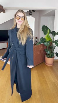 Abrigo largo de trench navy hecho de tela con un cinturón, botones de doble botonadura y dos bolsillos frontales. Llevado con pantalones negros en un espacio interior bien iluminado.