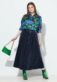 Bloemige overhemd met knopen in blauw, groen en zwart; marineblauwe geplisseerde maxi-rok; groene handtas; groene enkellaarsjes met glans.