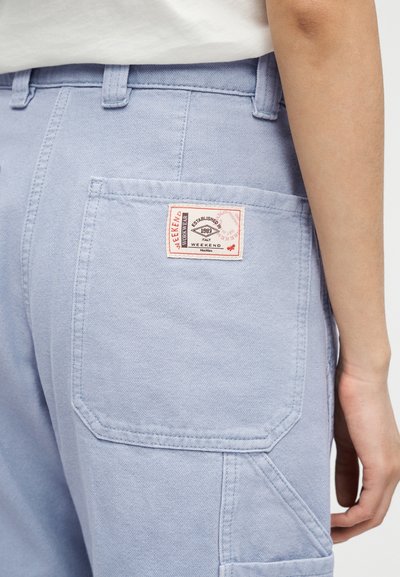 Ανοιχτό μπλε παντελόνι με πίσω τσέπη που διαθέτει ραμμένη ορθογώνια ετικέτα, φορεμένο από άτομο που στέκεται με το δεξί του χέρι ορατό.