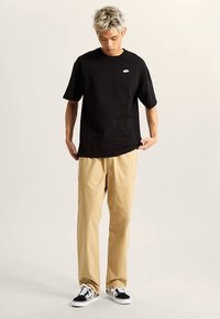 Czarny t-shirt z krótkim rękawem i okrągłym dekoltem z małym logo; zestawiony z beżowymi spodniami i czarno-białymi sneakersami w wzory.