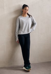Grå, kort sweatshirt, svarte tettsittende bukser, grønne joggesko med hvite såler. Modellen står mot en lys, teksturert vegg.