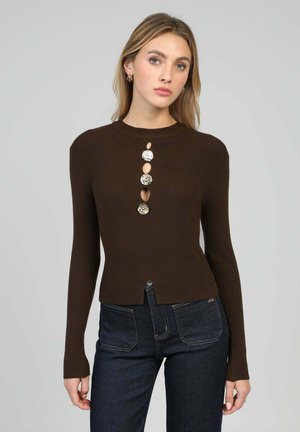 Pull marron côtelé à manches longues, avec un col rond et un détail de boutons décoratifs sur le devant. Associé à un jean en denim foncé.