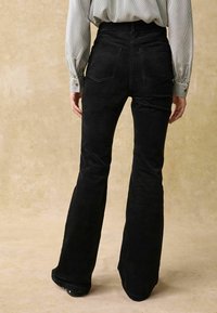 Pantalons évasés en velours côtelé noir à taille haute, avec deux poches au dos et une finition lisse et texturée. Le modèle porte une chemise rayée.