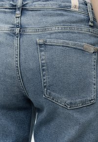 Κοντινό πλάνο της πίσω πλευράς ενός μπλε τζιν με ορατή τσέπη, θηλιά για ζώνη και επώνυμη ετικέτα στη ζώνη και την τσέπη.