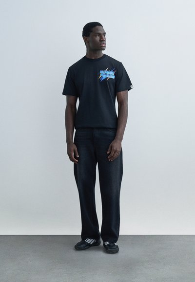 Άνδρας που φοράει μαύρο μπλουζάκι με τύπωμα, φαρδιά μαύρη παντελόνα και μαύρα αθλητικά παπούτσια Adidas με λευκές ρίγες, στέκεται μπροστά σε ένα απλό ανοιχτό γκρι τοίχο.