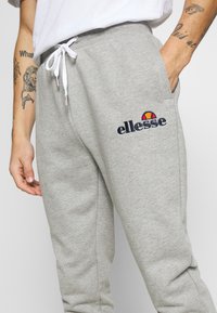 Grå sweatpants i blødt materiale, med en hvid snor og et broderet "ellesse" logo med farverige accenter på låret.