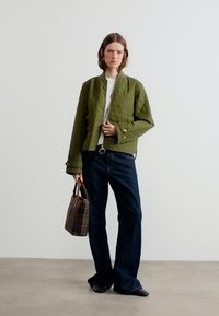 Γυναίκα με κιμονό σε χακί πράσινο χρώμα, λευκό πουκάμισο, σκούρα τζιν με φαρδιά μπατζάκια, μαύρα παπούτσια, κρατώντας καρό καφέ τσάντα, στέκεται σε απλό φόντο.