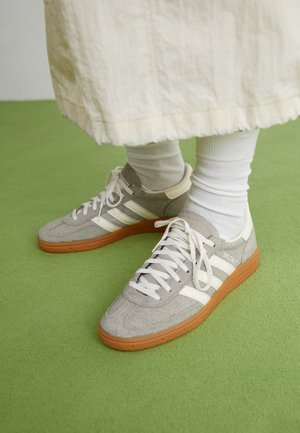 Fødder iført grå ruskinds Adidas-sneakers med hvide striber og gummisåler, kombineret med hvide sokker og en beige nederdel på grøn gulv.